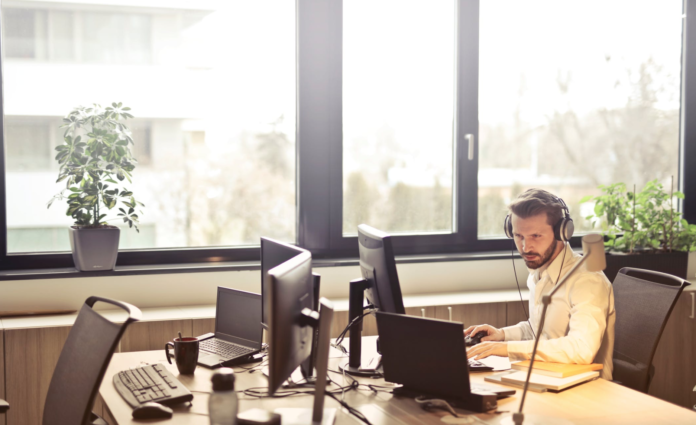 man in office inn front of window working on laptop wearing headphones